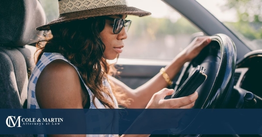 Woman in hat and glasses looking at phone behind steering wheel.
